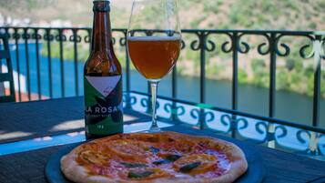 Una botella de cerveza IPA La Rosa, un vaso de cerveza y una pizza sobre una mesa con vistas a un río y colinas.