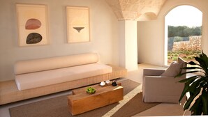 Una sala de estar moderna con un sofá, una mesa de centro de madera y una silla. Hay dos cuadros enmarcados en la pared y vistas al exterior a través de una ventana arqueada.