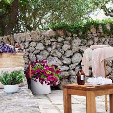 A spa area with a stone wall, a wooden table with a bottle, towels, and potted plants.