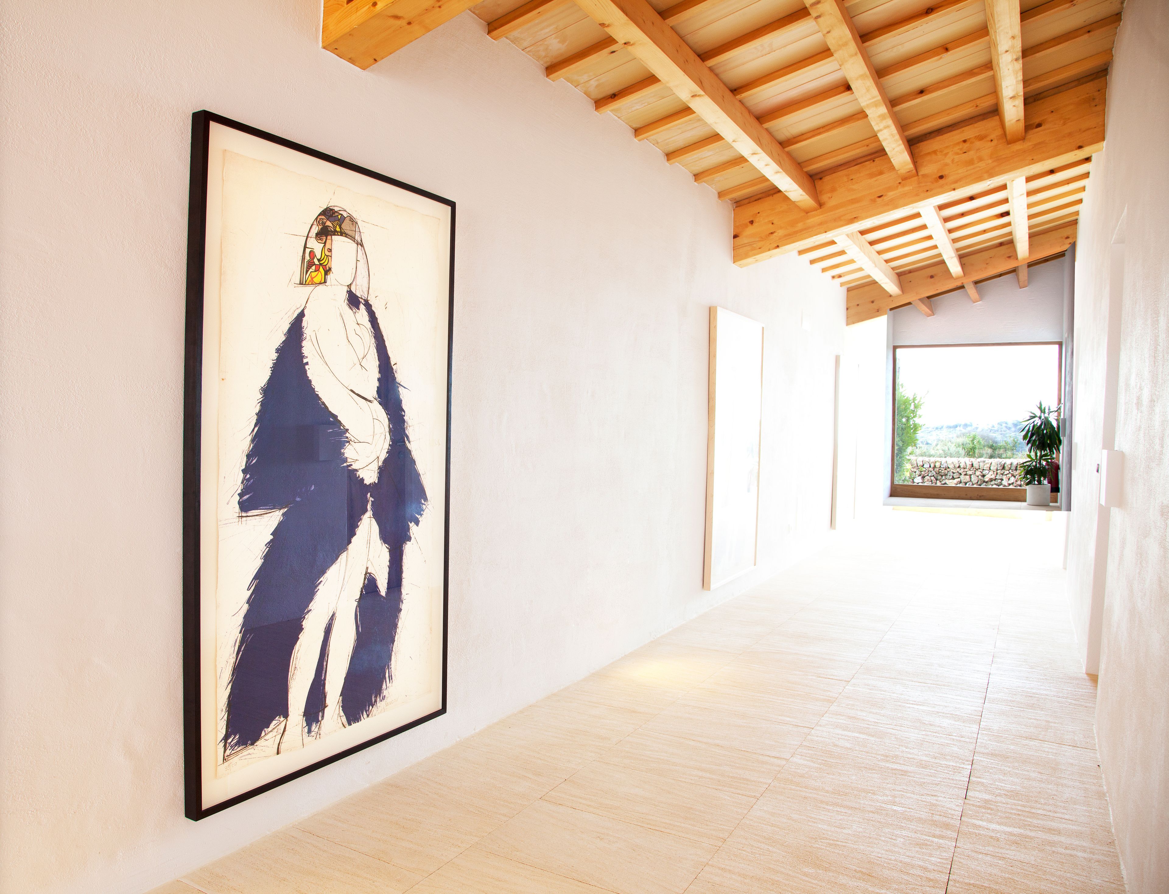 A hallway with a framed abstract painting on the left, wooden beams on the ceiling, and a view of a garden through a window on the right.