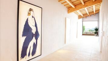 A hallway with a framed abstract painting on the left, wooden beams on the ceiling, and a view of a garden through a window on the right.
