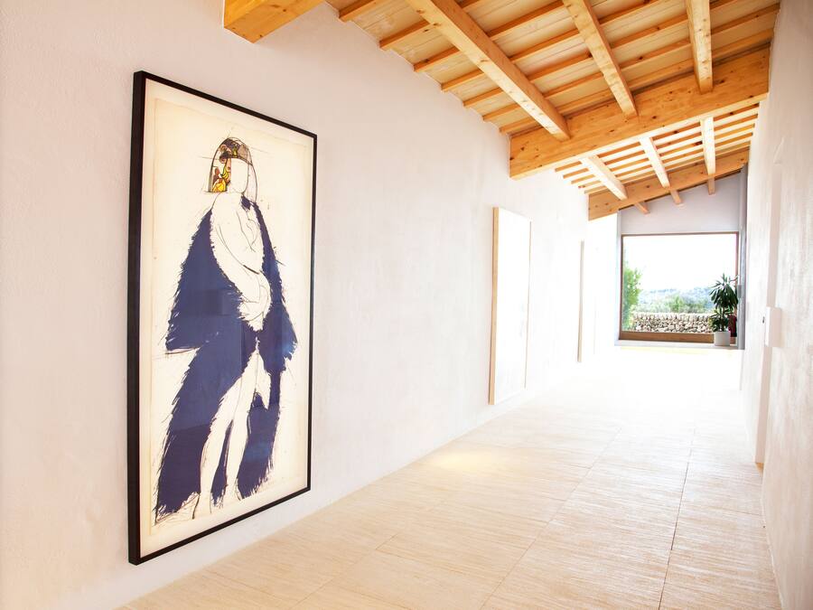 A hallway with a framed abstract painting on the left, wooden beams on the ceiling, and a view of a garden through a window on the right.