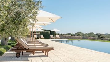 A poolside area with lounge chairs and umbrellas, a stone wall, and a clear sky.