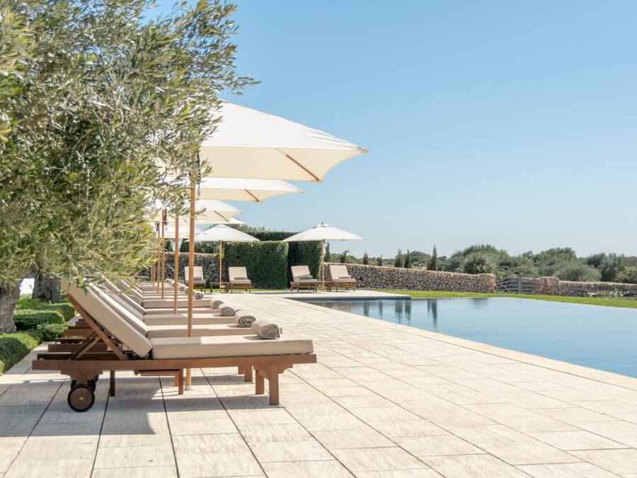 A poolside area with lounge chairs and umbrellas, a stone wall, and a clear sky.