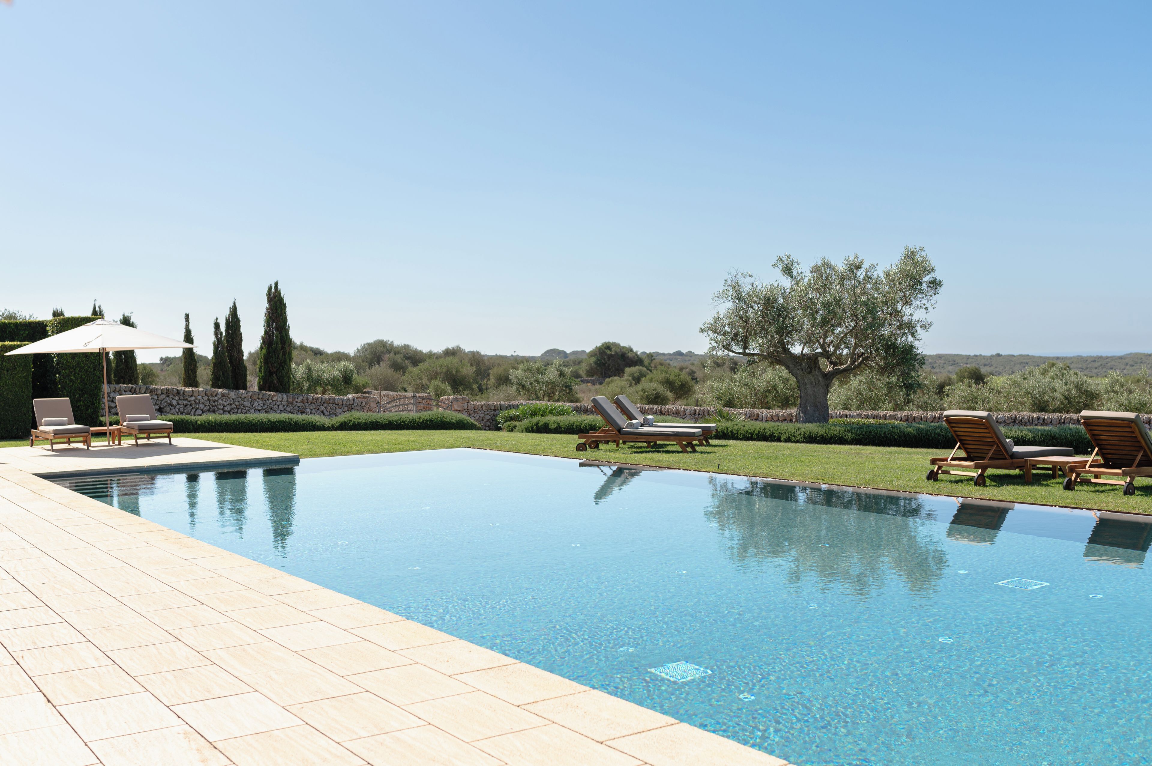 A rectangular swimming pool with a tiled deck, surrounded by lounge chairs and a stone wall.