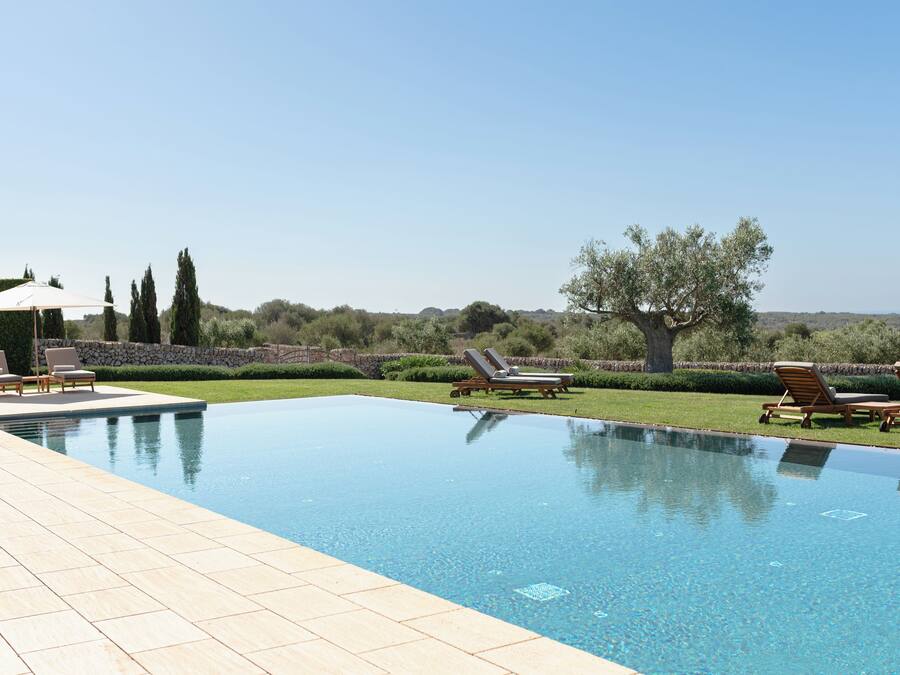 A rectangular swimming pool with a tiled deck, surrounded by lounge chairs and a stone wall.