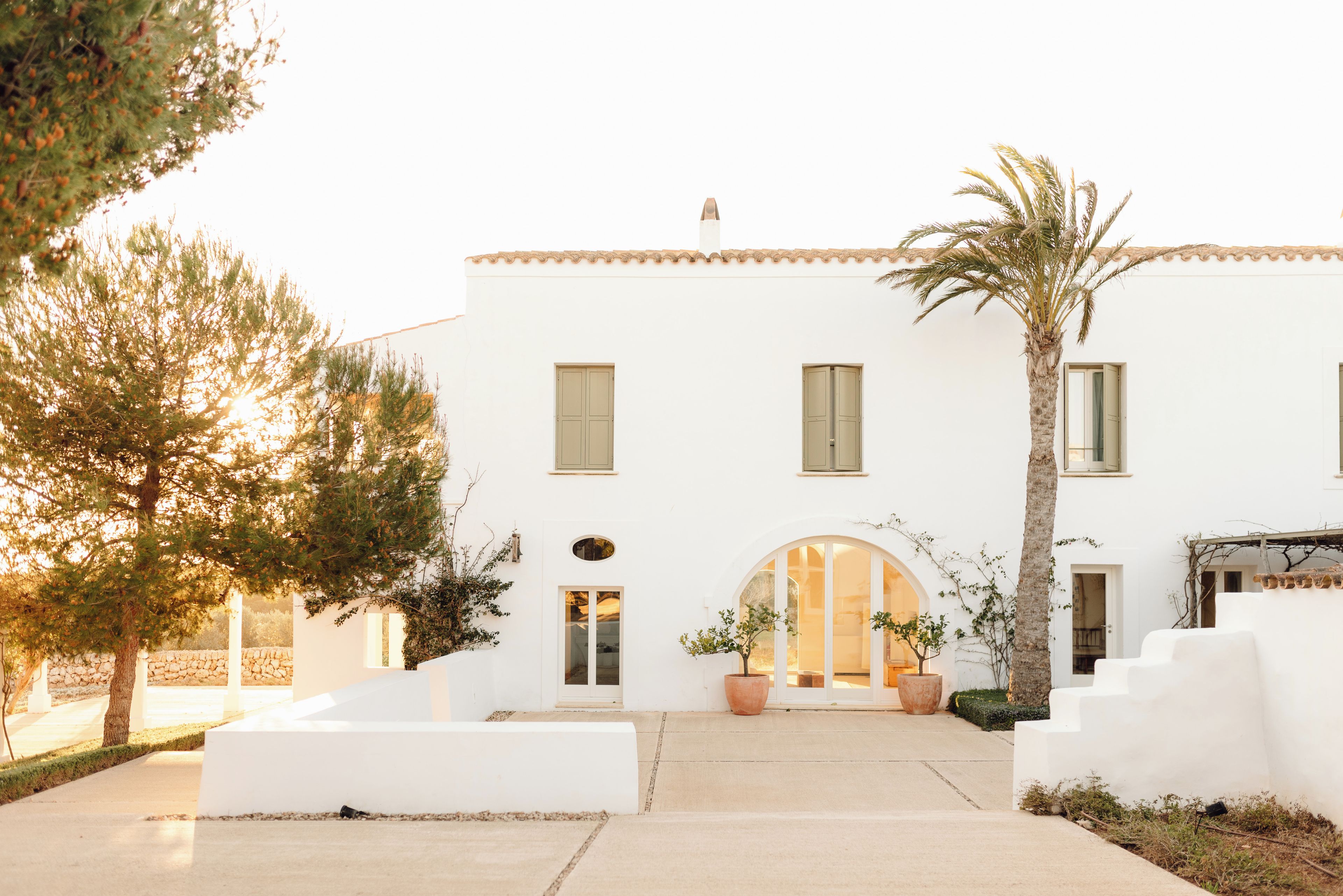 A white building with a central entrance, two windows on the upper floor, and a palm tree to the right.