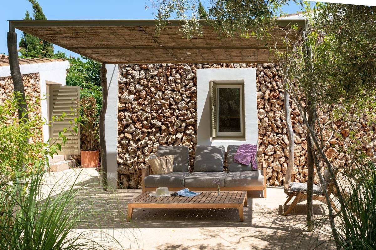 A covered outdoor seating area with a stone wall backdrop, a wooden table, and a sofa with cushions.