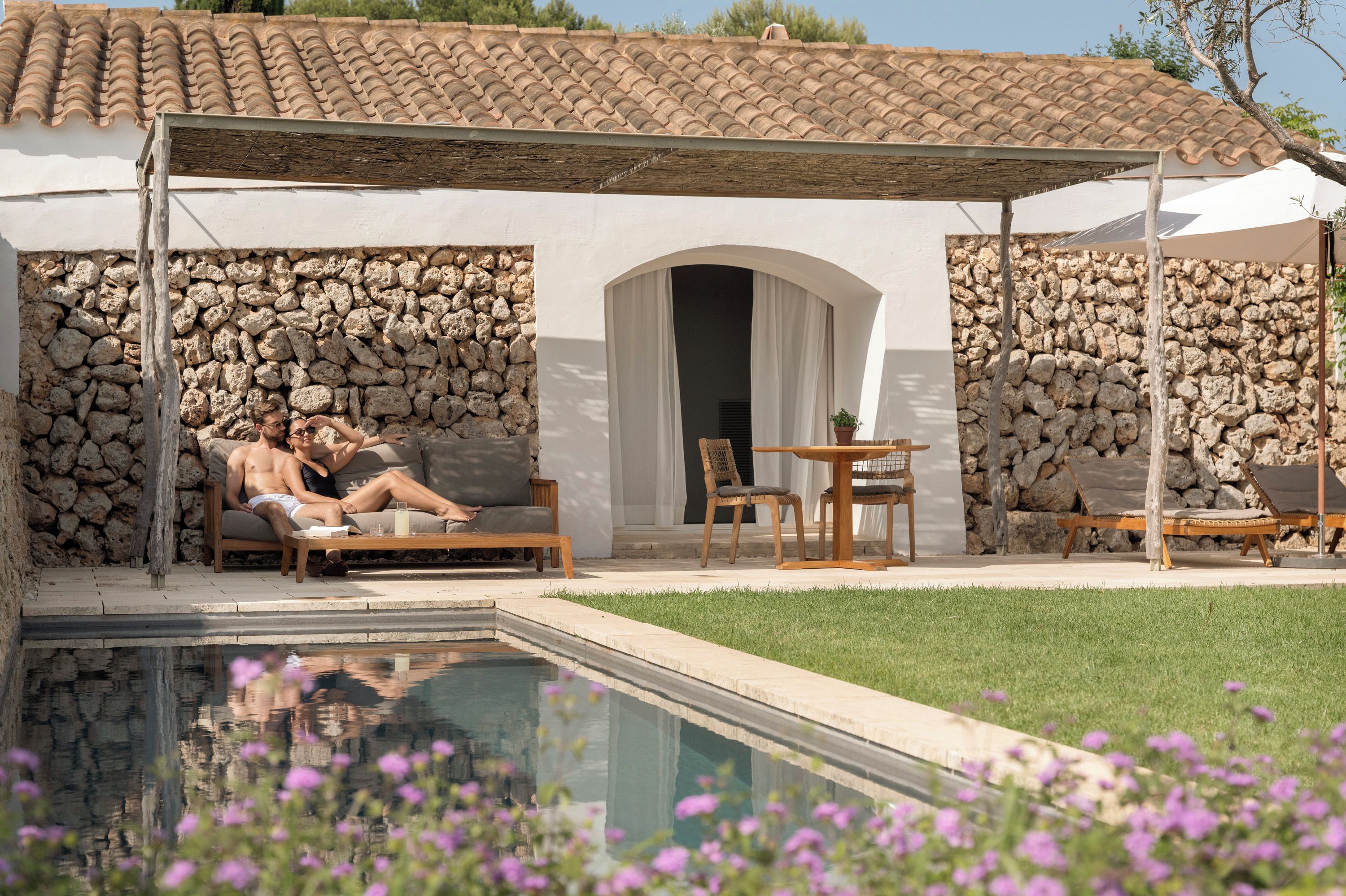 Zona junto a la piscina con muro de piedra, espacio de descanso cubierto y vista despejada a la piscina.