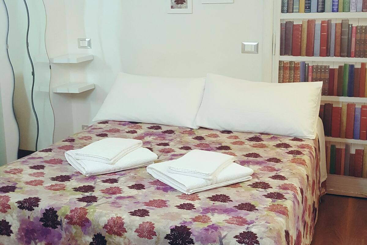 Ein ordentlich bezogenes Bett mit floralem Überwurf, ein Nachttisch mit Lampe und eine Bücherwand voller Bücher.