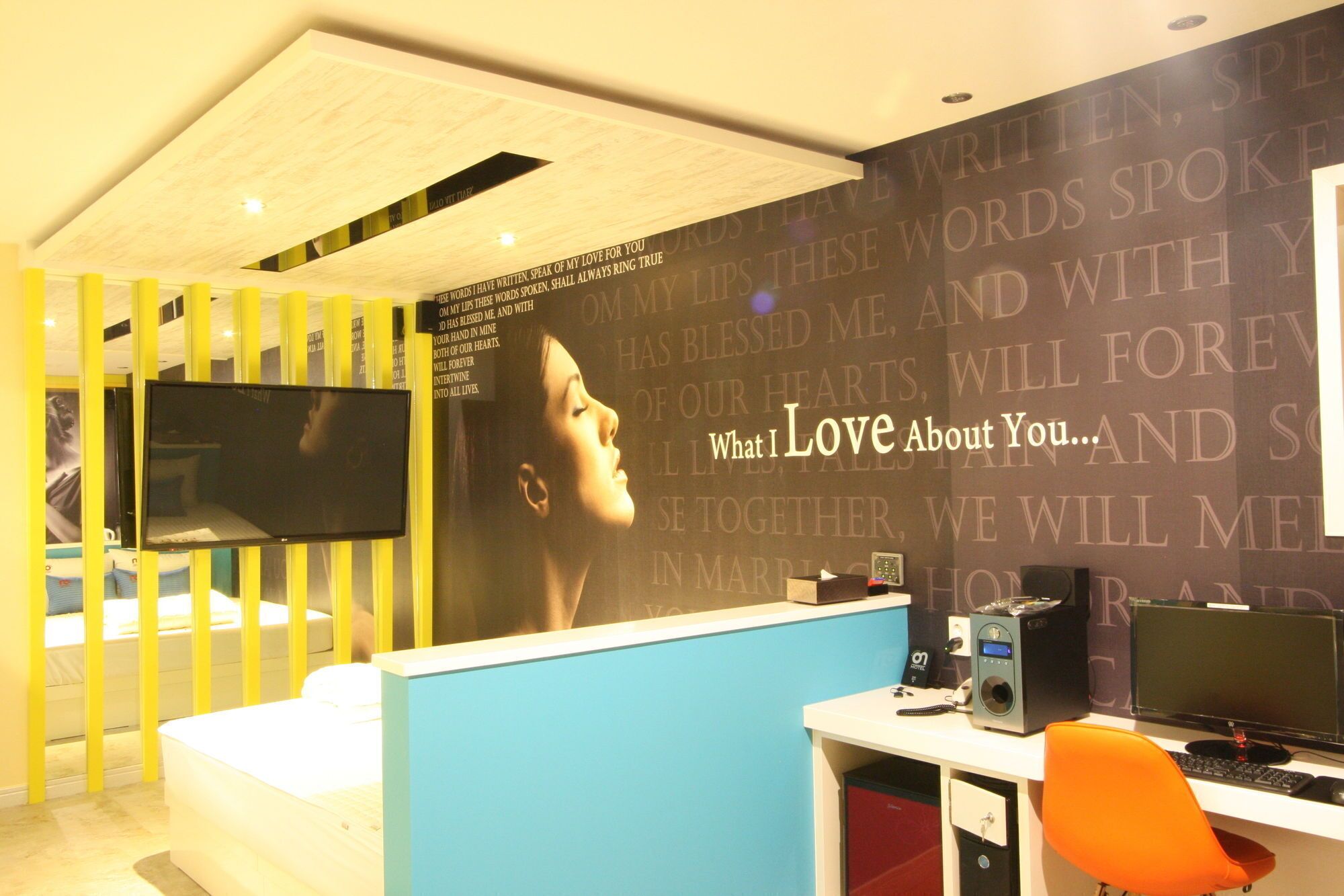 A modern hotel room with a large wall mural, a flat-screen TV, a desk with a computer, and a comfortable bed.