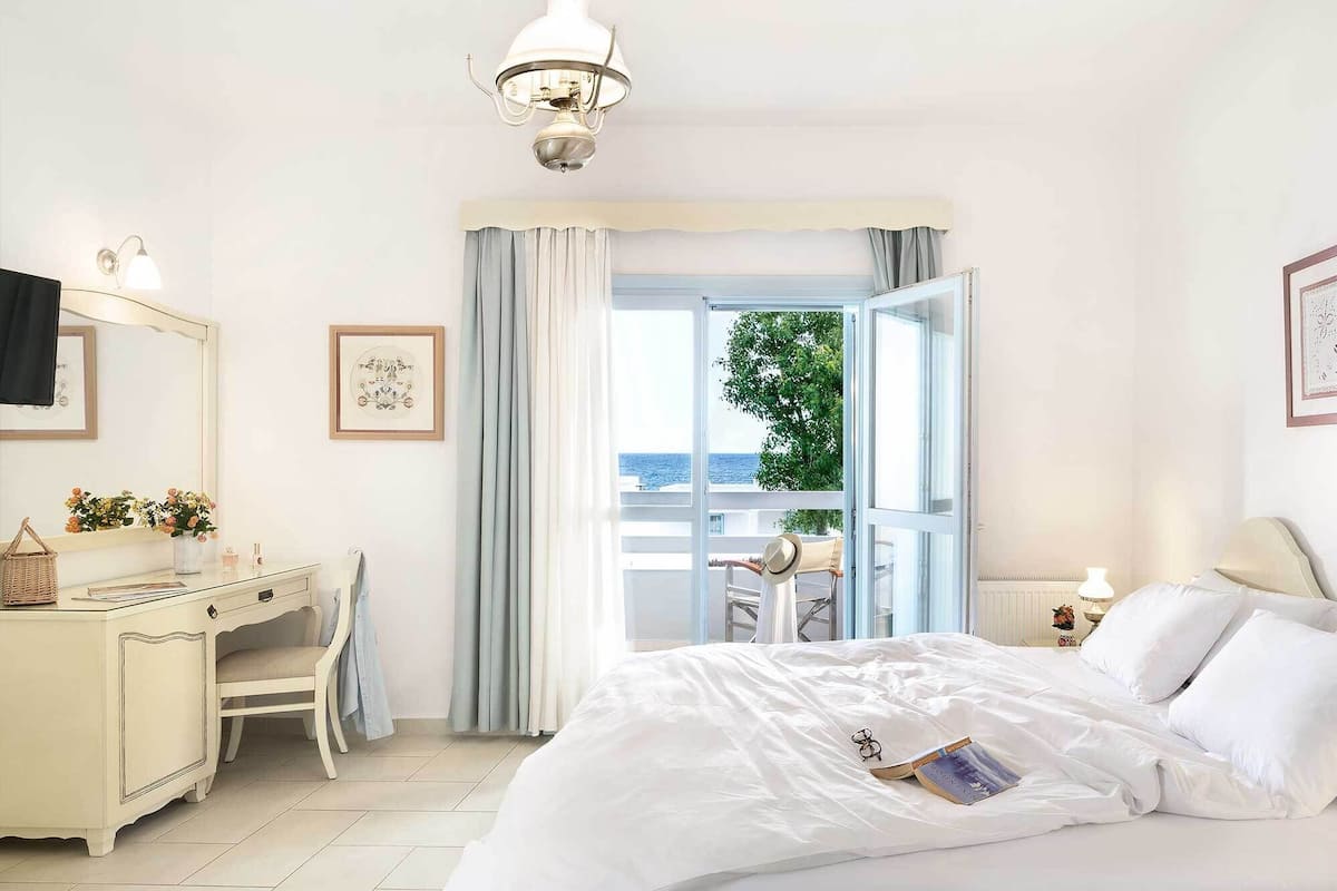 A bedroom with a bed, a desk, a chair, a dresser, and a view of the sea through an open door.