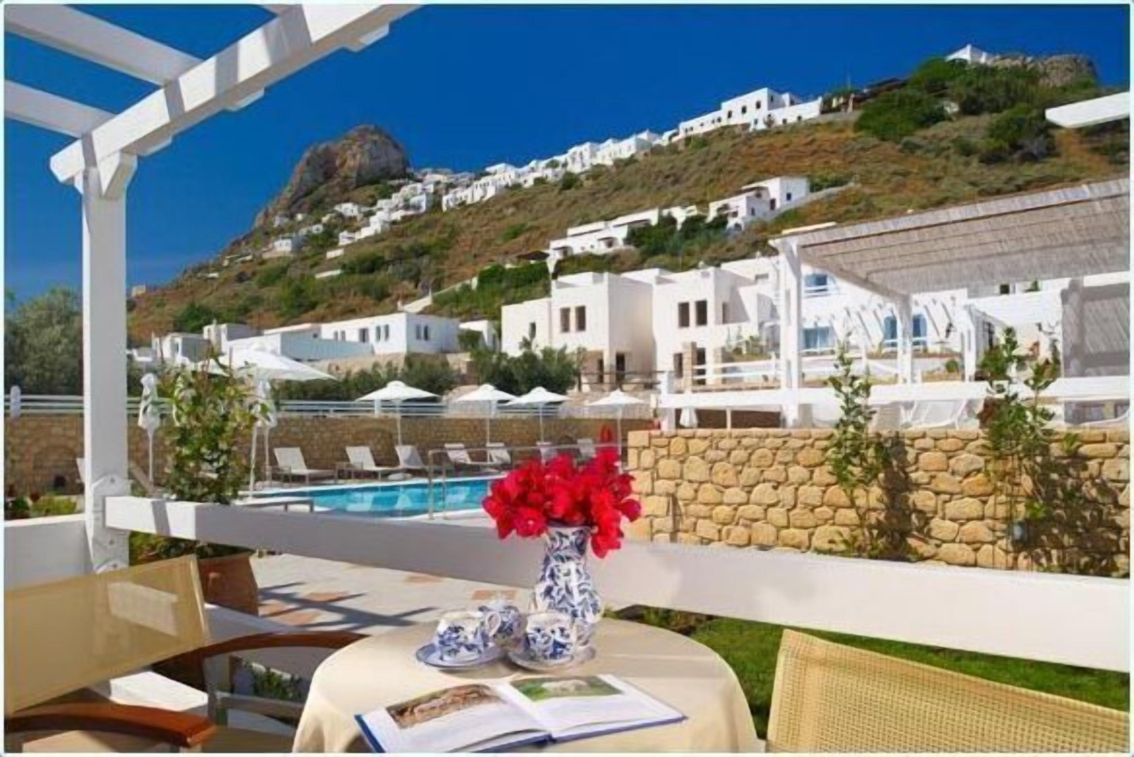 A terrace with a pool, white buildings in the background, and a stone wall.