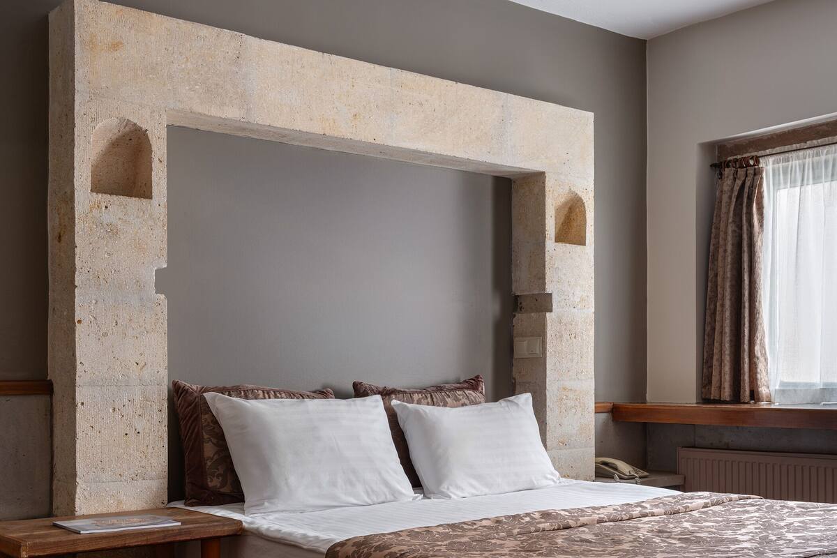 A bedroom with a stone headboard, a bed with white and brown bedding, a wooden nightstand, a red and black striped rug, and a window with curtains.