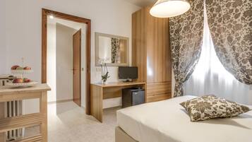 Habitación de hotel con cama, escritorio, televisor, refrigerador pequeño, espejo y ventana con cortinas.