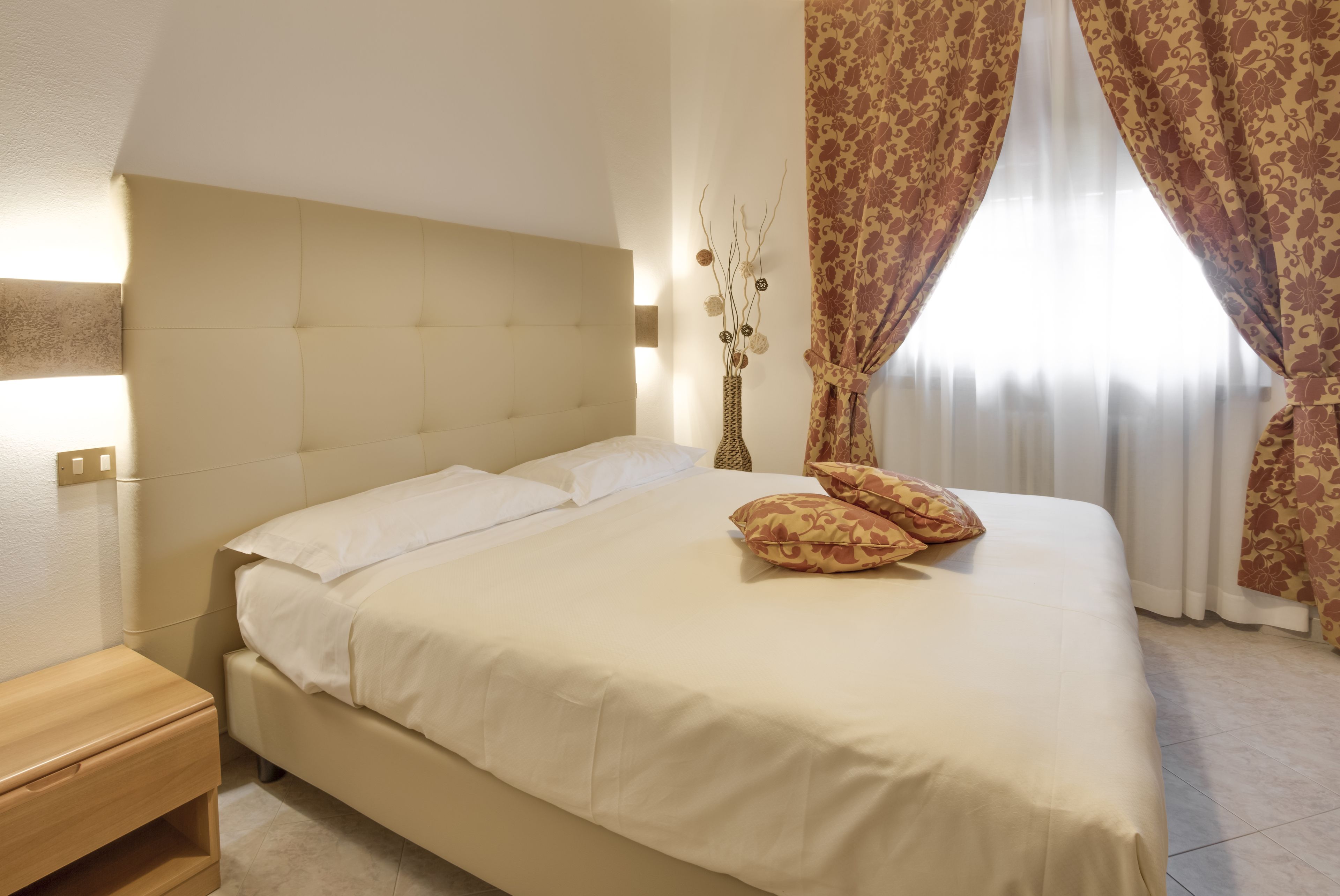 Un letto rifatto con lenzuola bianche e due cuscini fantasia, un comodino in legno e un vaso con dei rami sul comodino.