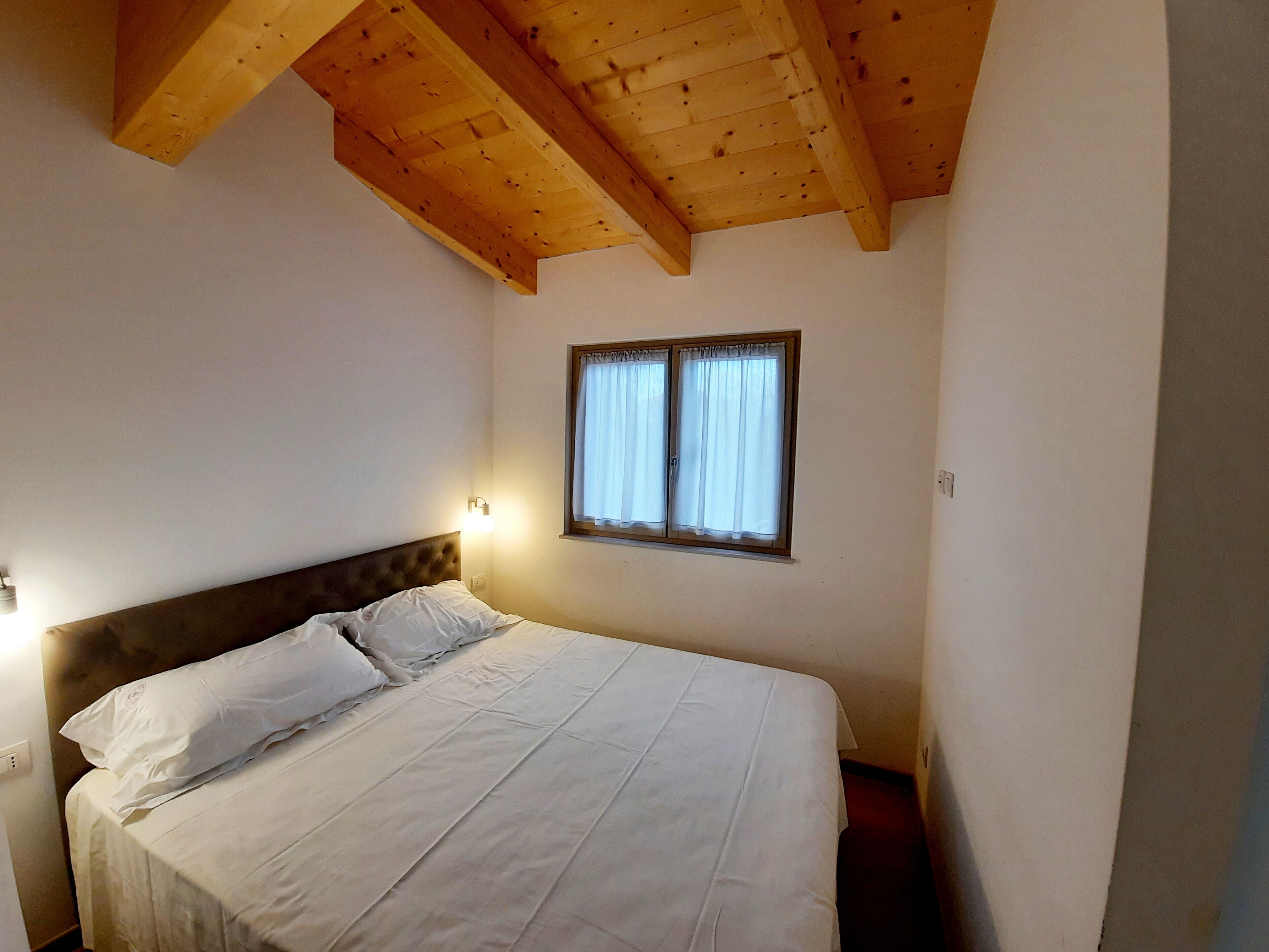 A bedroom with a wooden ceiling, a single bed with white bedding, a nightstand, and a window with curtains.