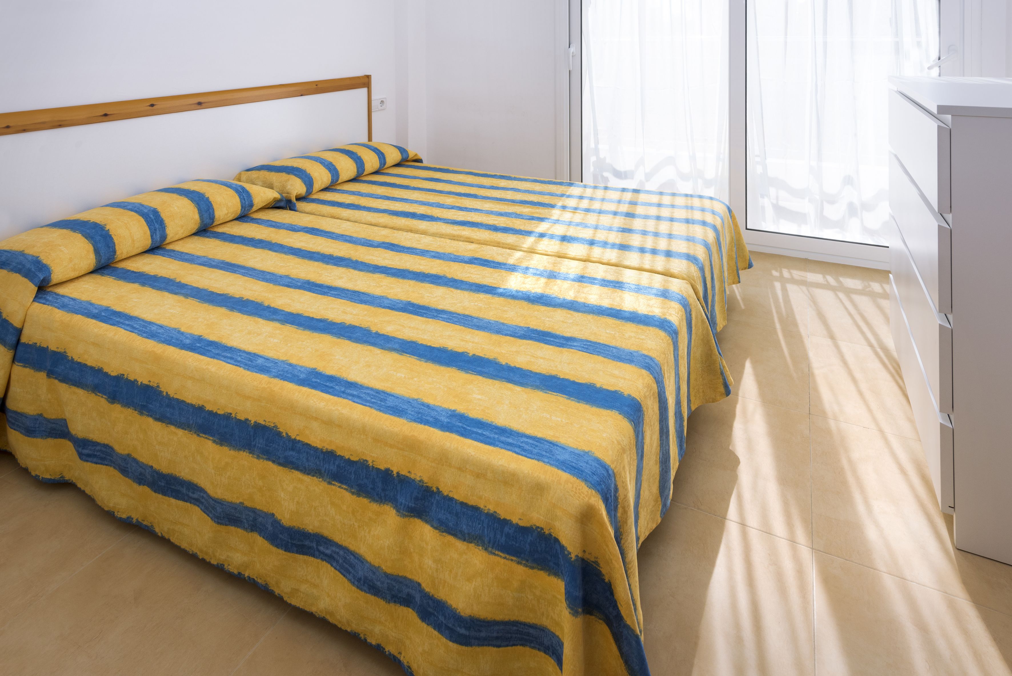 Una habitación con una cama cubierta con ropa de cama a rayas, un cabecero de madera, un armario blanco y un ventanal con cortinas traslúcidas.