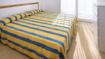Una habitación con una cama cubierta con ropa de cama a rayas, un cabecero de madera, un armario blanco y un ventanal con cortinas traslúcidas.