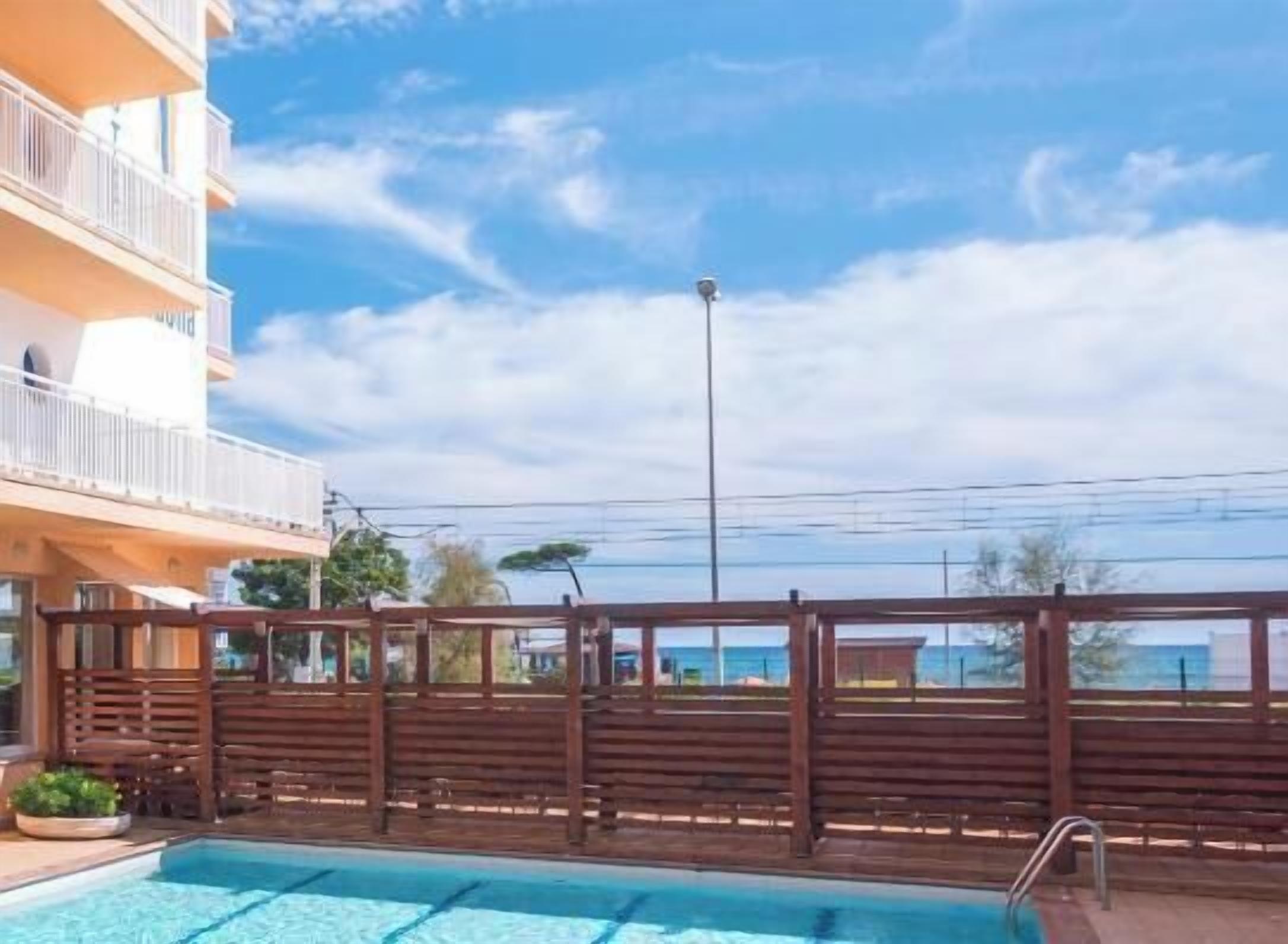 A swimming pool with a wooden fence, a clear sky, and a building with balconies.