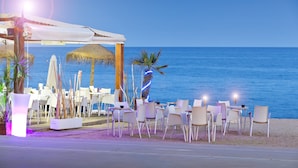 Un restaurant situé en bord de mer, avec une terrasse, un parasol en chaume et une vue sur l’océan.