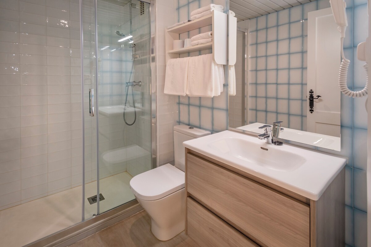 Een moderne badkamer met een douchecabine van glas, een wit toilet, een wastafelmeubel met spiegel en een handdoekrek met netjes opgevouwen handdoeken.