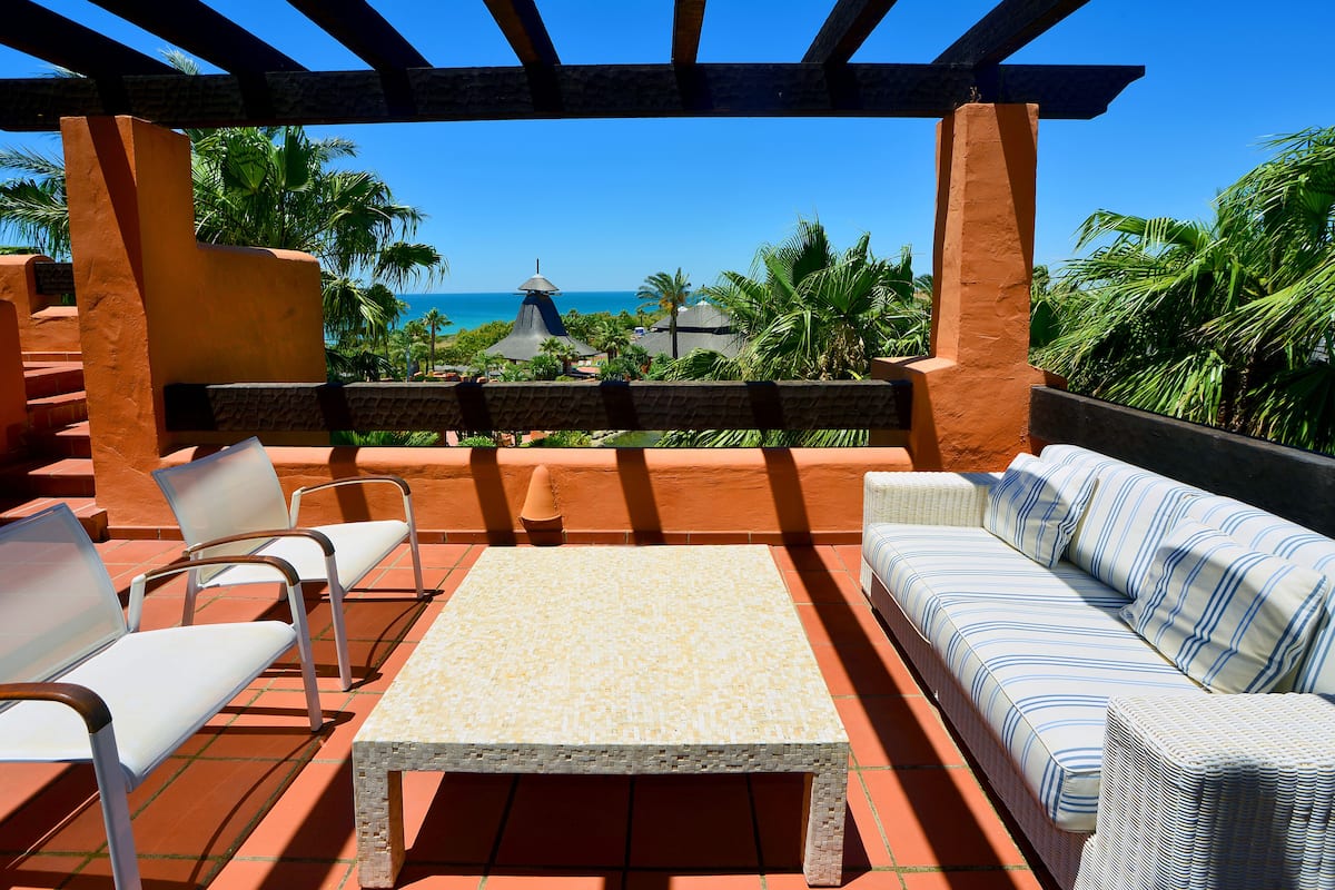 A covered outdoor seating area with a view of the ocean, featuring a sofa, chairs, and a table.