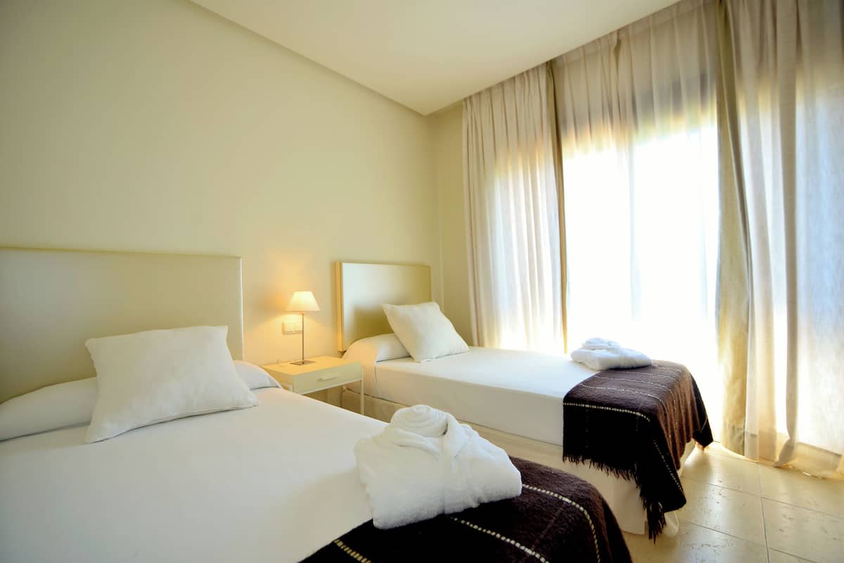 A hotel room with two beds, a nightstand with a lamp, and a window with curtains.