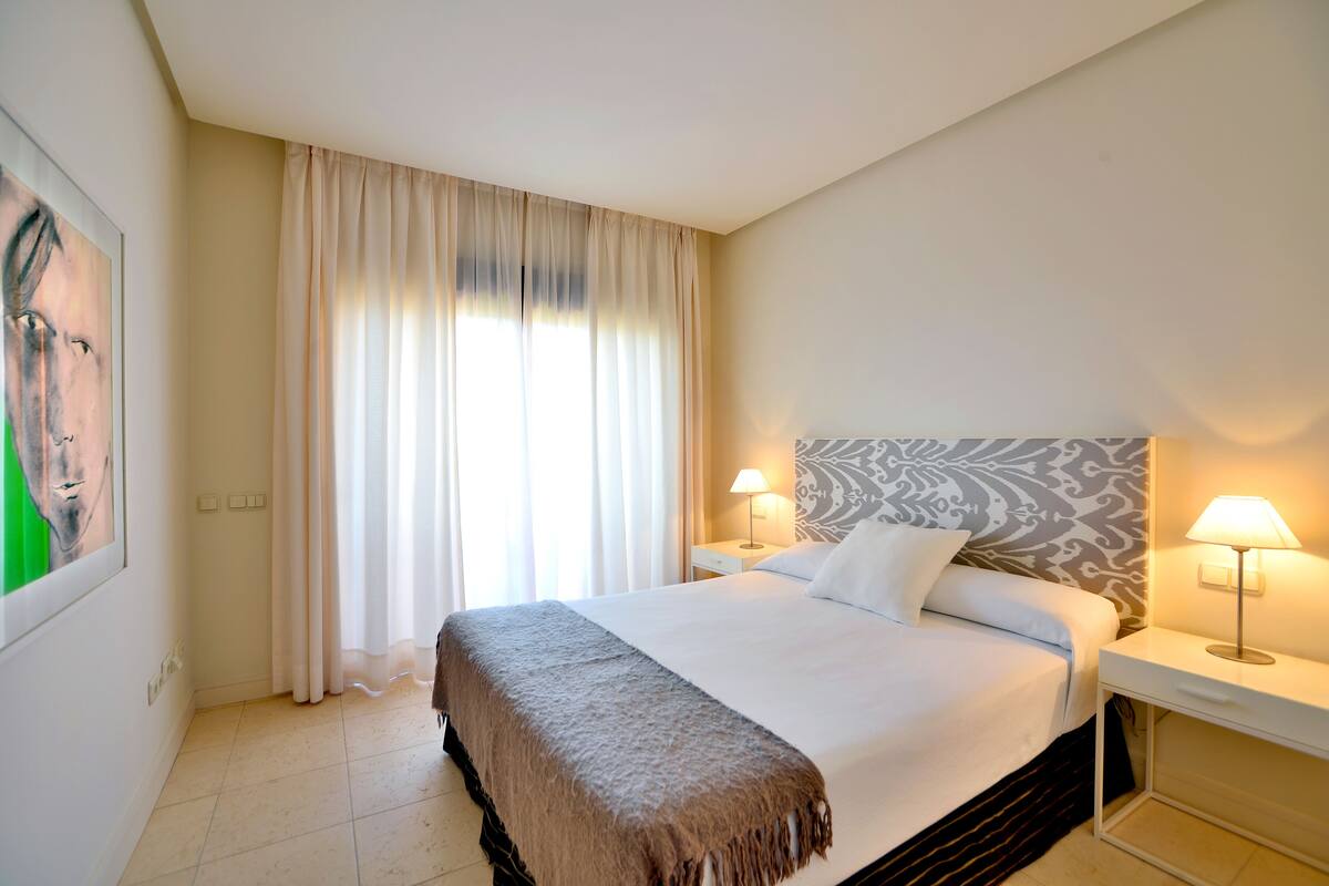 A hotel room with a bed, two bedside lamps, a nightstand, a large window with curtains, and a painting on the wall.
