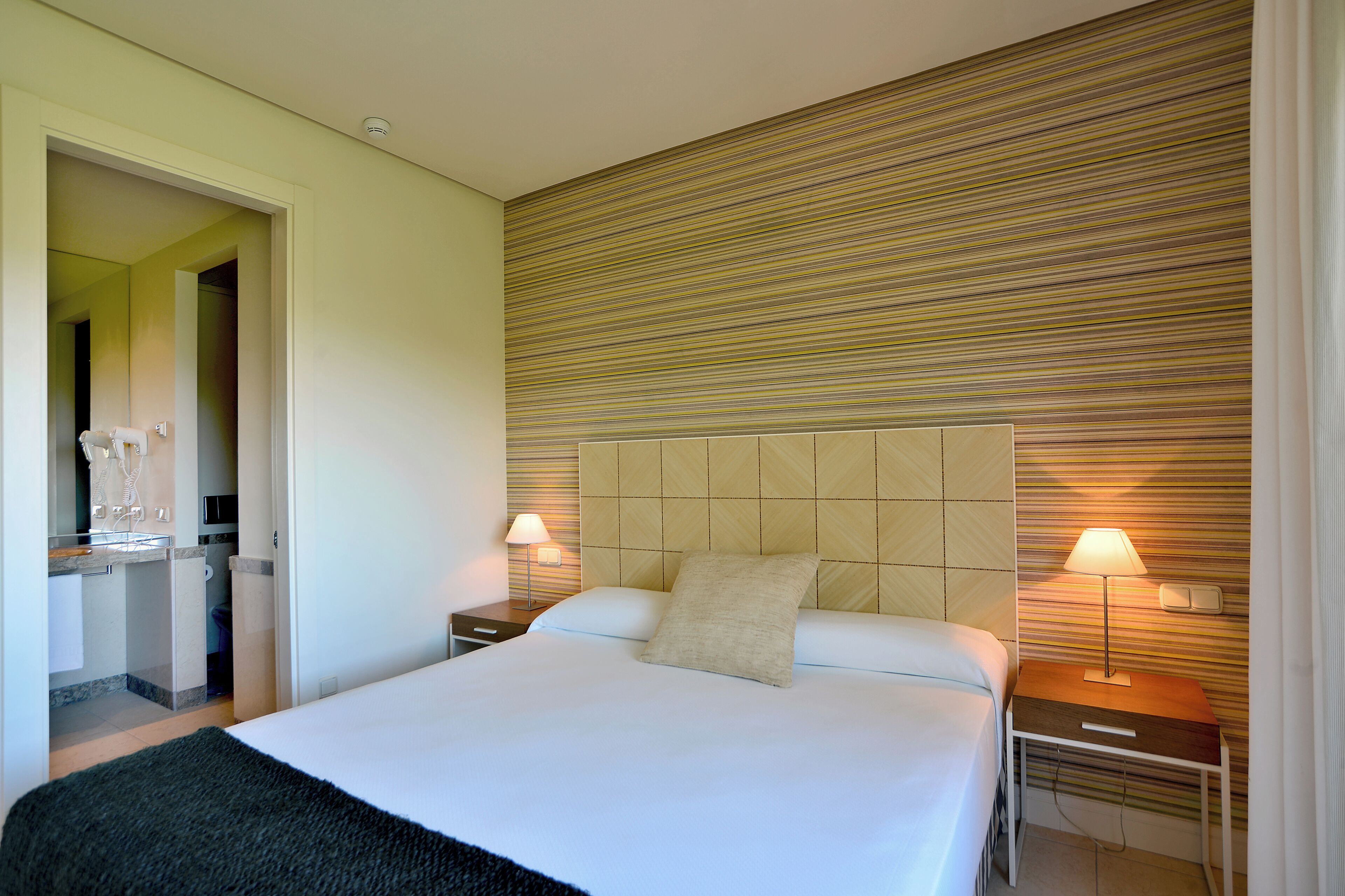 Ein Hotelzimmer mit einem Bett, zwei Nachttischen, einer Lampe, einer Wand mit Streifenmuster und einem durchgegangenen Tür sichtbaren Badezimmer.