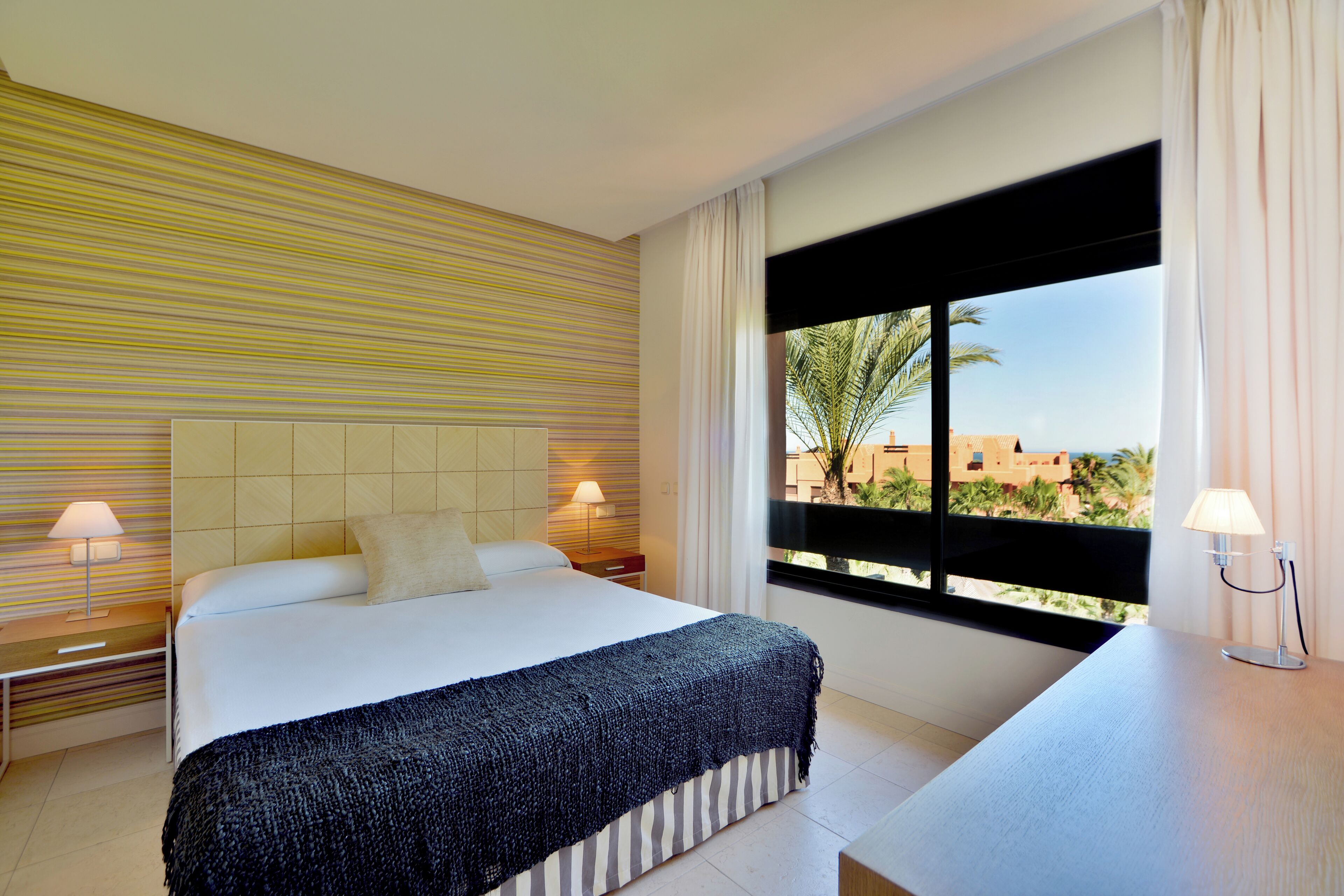 Ein Hotelzimmer mit einem großen Bett, einem Nachttisch, einem Schreibtisch und einem Fenster mit Blick auf Palmen und Gebäude.