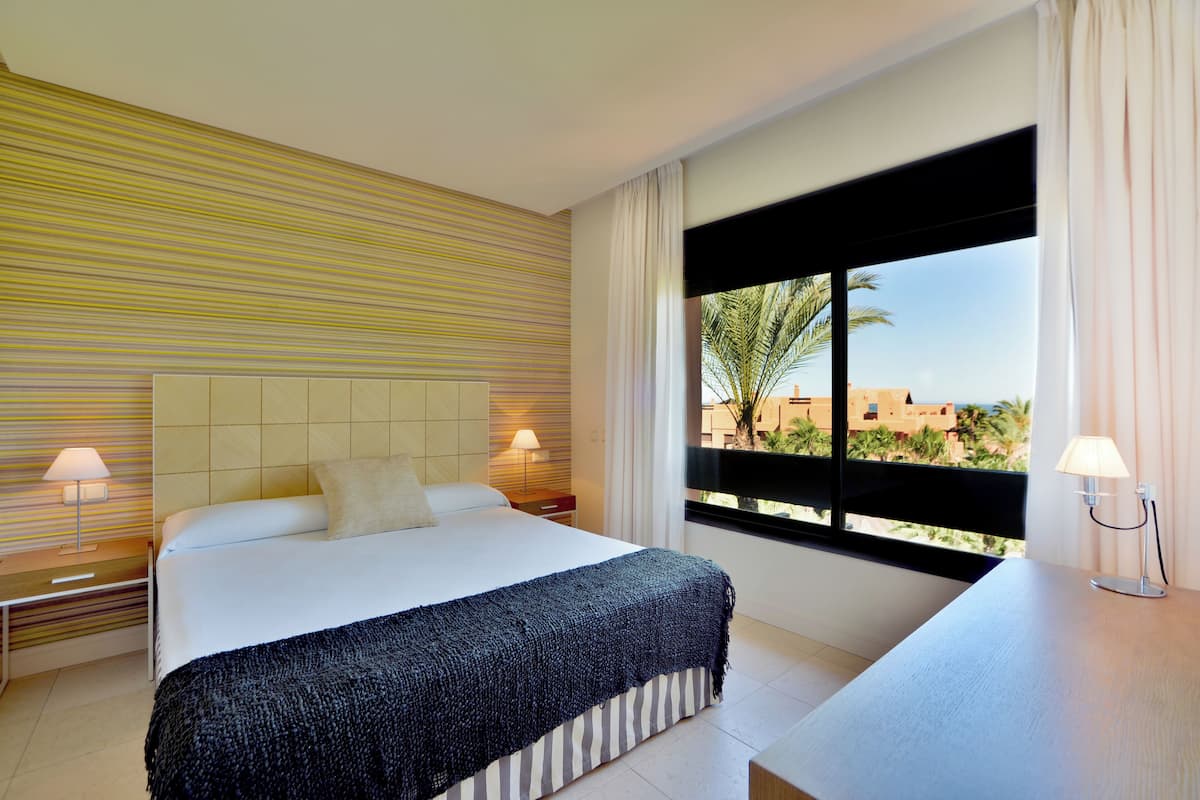 Habitación de hotel con una cama grande, una mesita de noche, un escritorio y una ventana con vistas a palmeras y edificios.