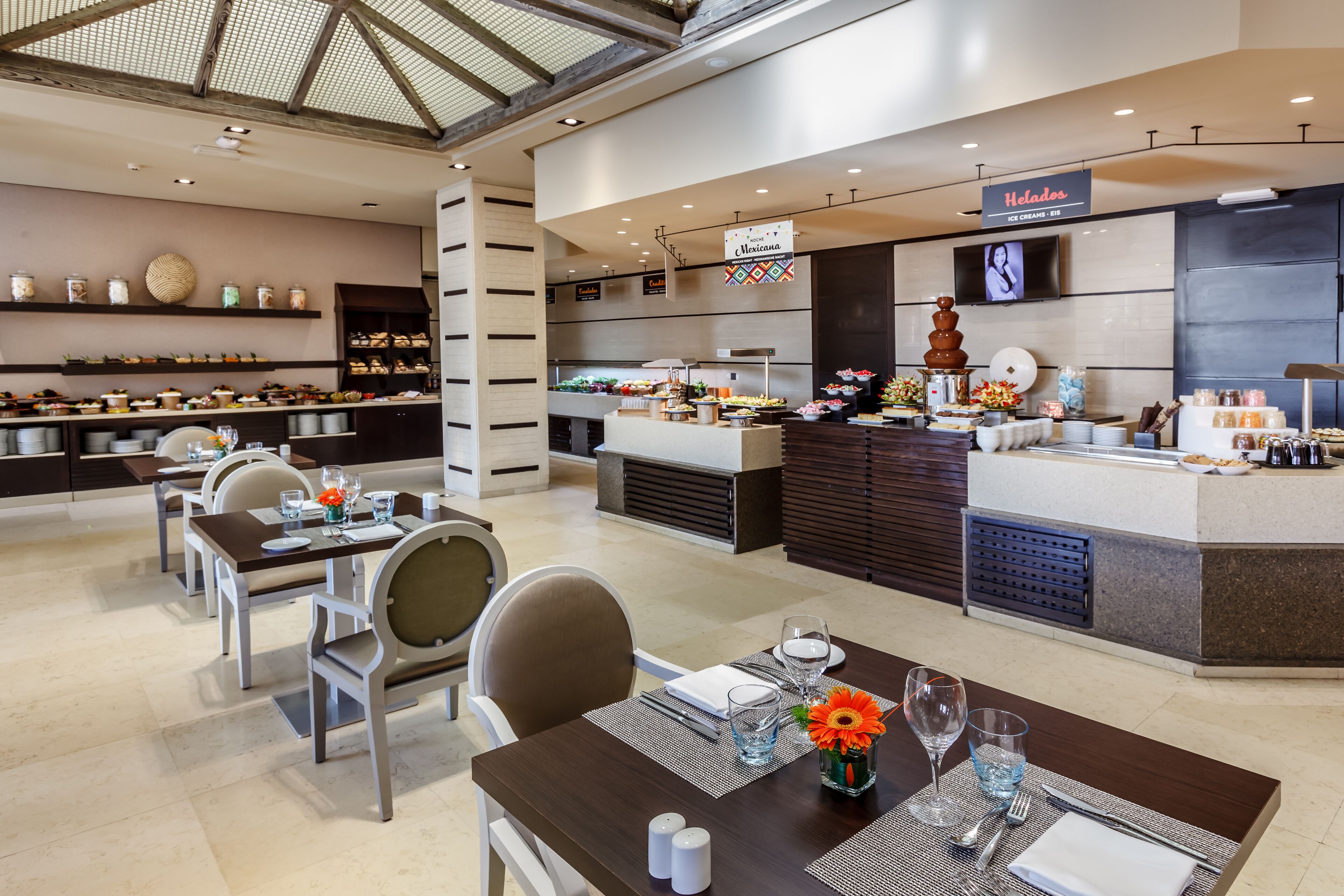 A modern restaurant with a central counter, multiple dining tables, and a variety of food displays.
