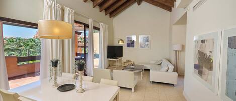 A spacious living area with a dining table, sofa, and TV. There is a balcony with plants and a view of greenery.