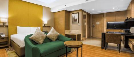 A hotel room with a bed, a green sofa, a small round table, a desk, and a kitchenette.