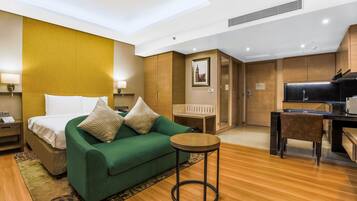 Une chambre d’hôtel comprenant un lit, un canapé vert, une petite table ronde, un bureau en bois et une kitchenette.