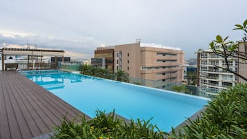 A rooftop swimming pool with a wooden deck, surrounded by greenery and overlooking a cityscape.