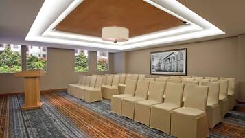 Un salón de conferencias con un atril de madera, hileras de sillas color beige, una alfombra estampada y ventanas amplias con vistas a zonas verdes.