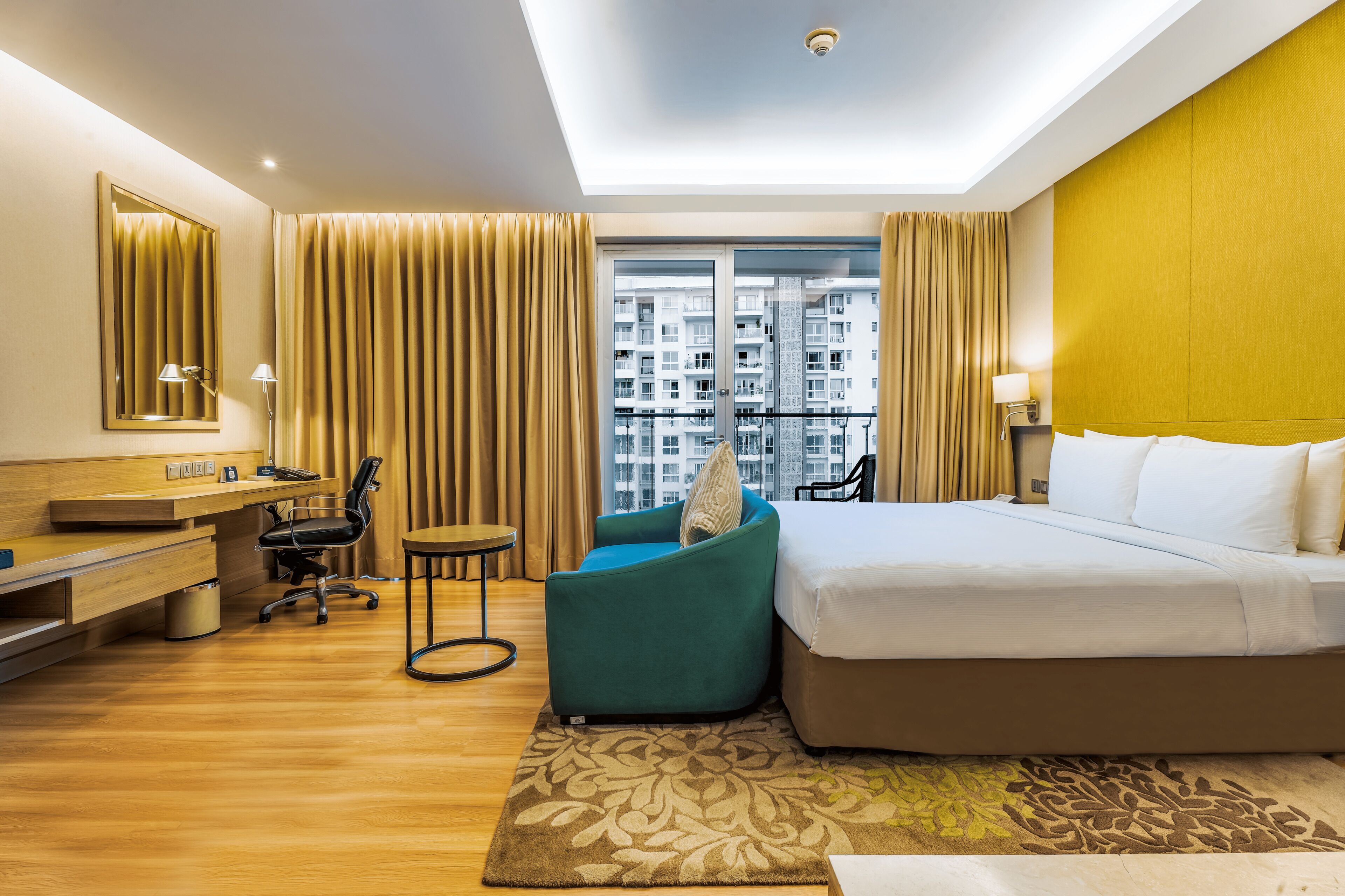 A modern hotel room with a large bed, a desk with a chair, a sofa, and a view of high-rise buildings.