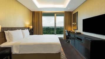 A hotel room with a large bed, a desk, a flat-screen TV, and a balcony with a view of greenery.