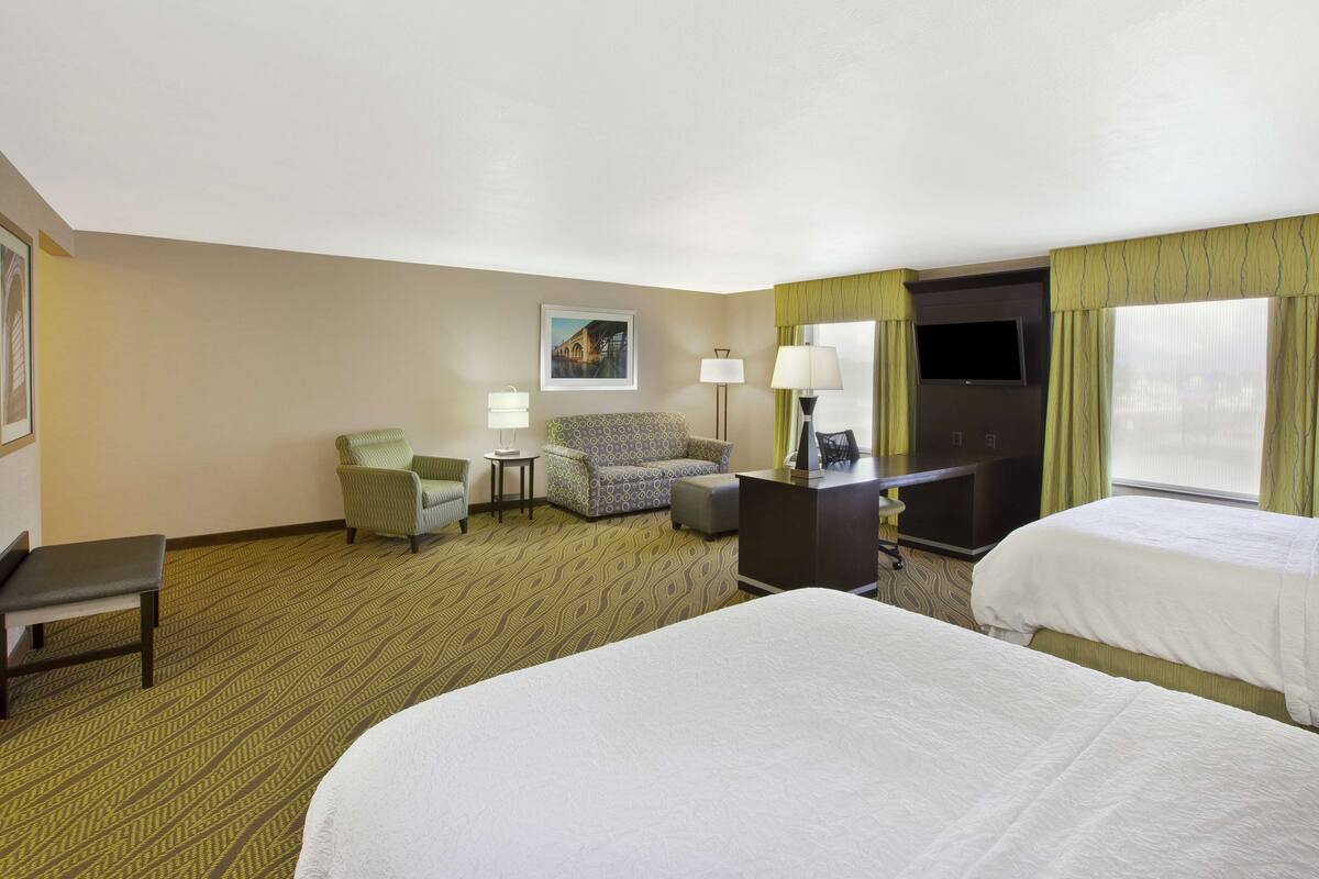 A hotel room with two beds, a desk, a chair, a small table, and a television.