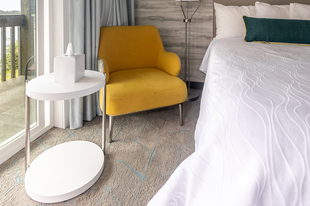 A hotel room with a bed, a yellow armchair, a round table, a lamp, and a view of the outdoors.