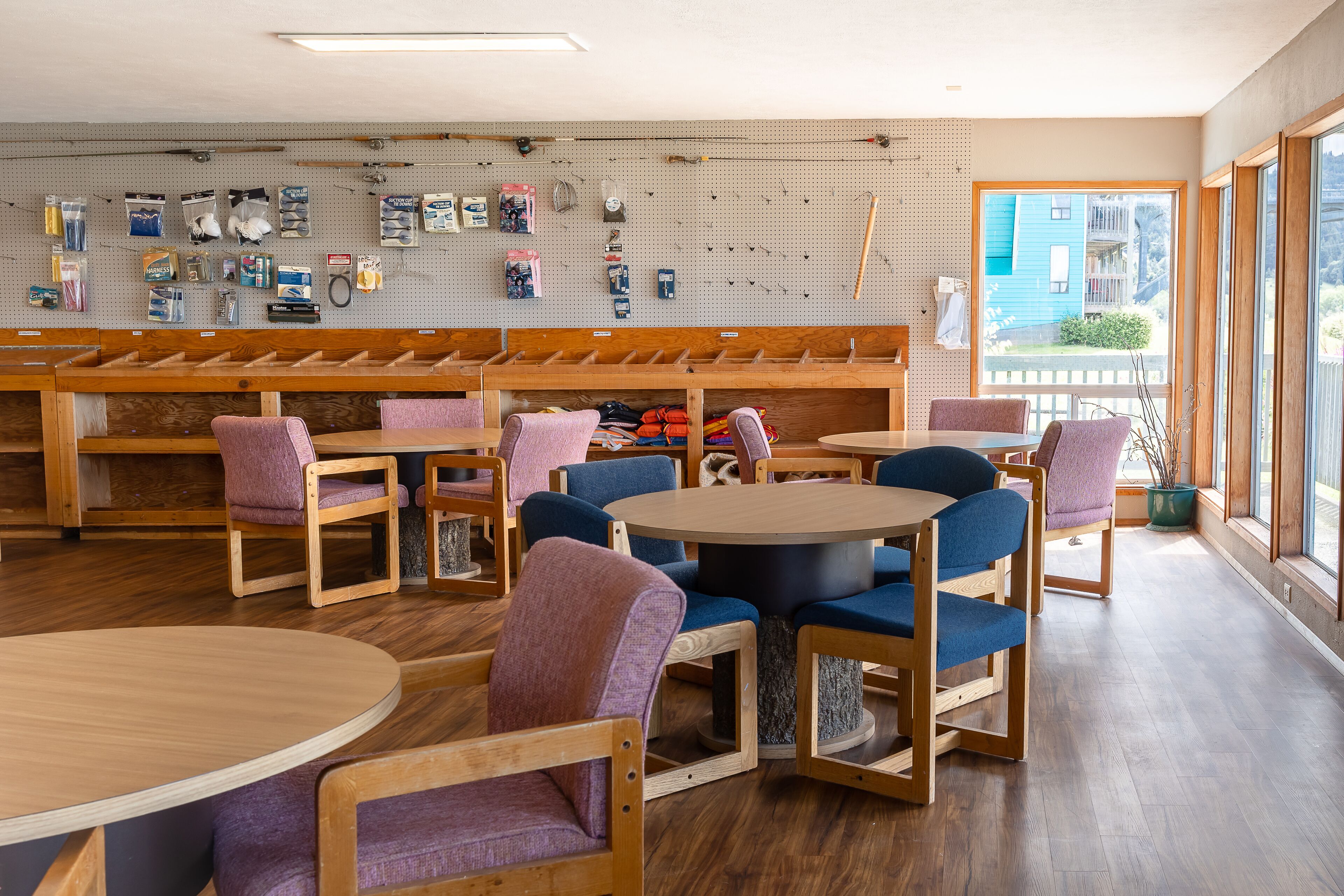 A restaurant with wooden tables and chairs, a pegboard wall with various items, and a view of a blue building outside.