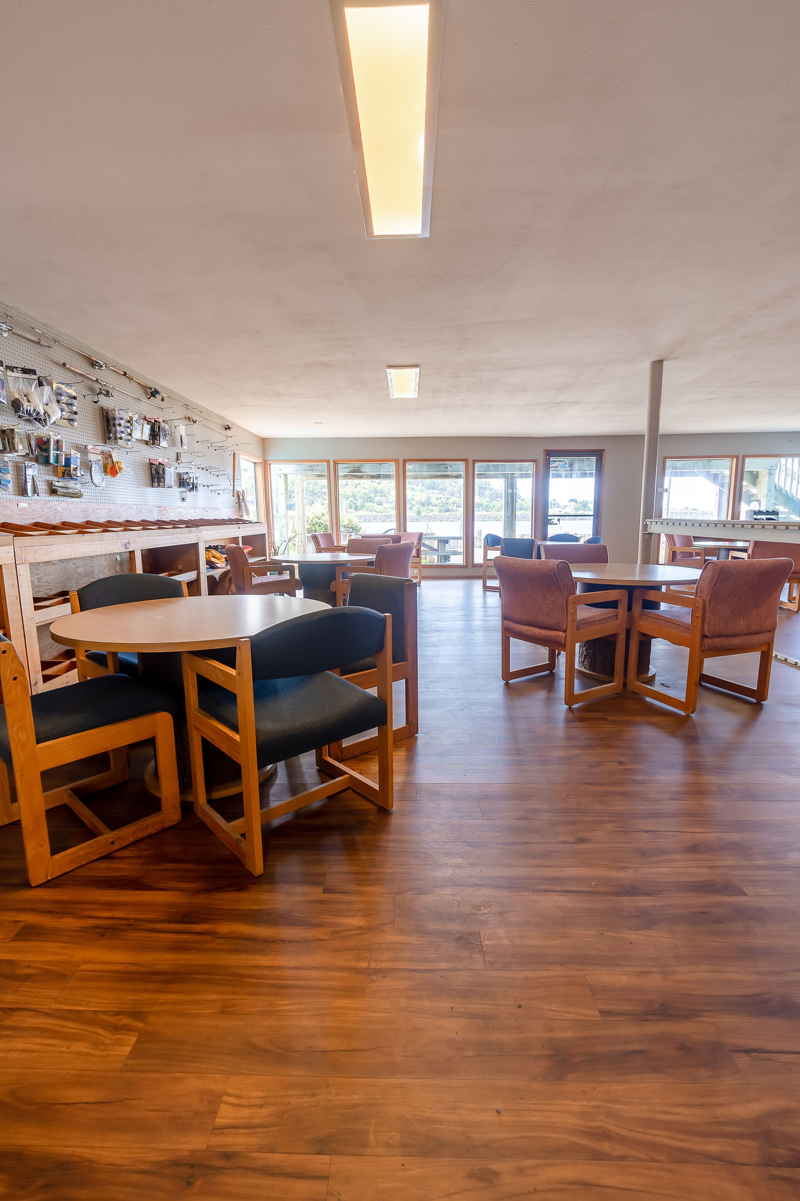 Een ruime eetruimte met houten tafels en stoelen, grote ramen die natuurlijk licht binnenlaten, en een gereedschapskast aan de muur.