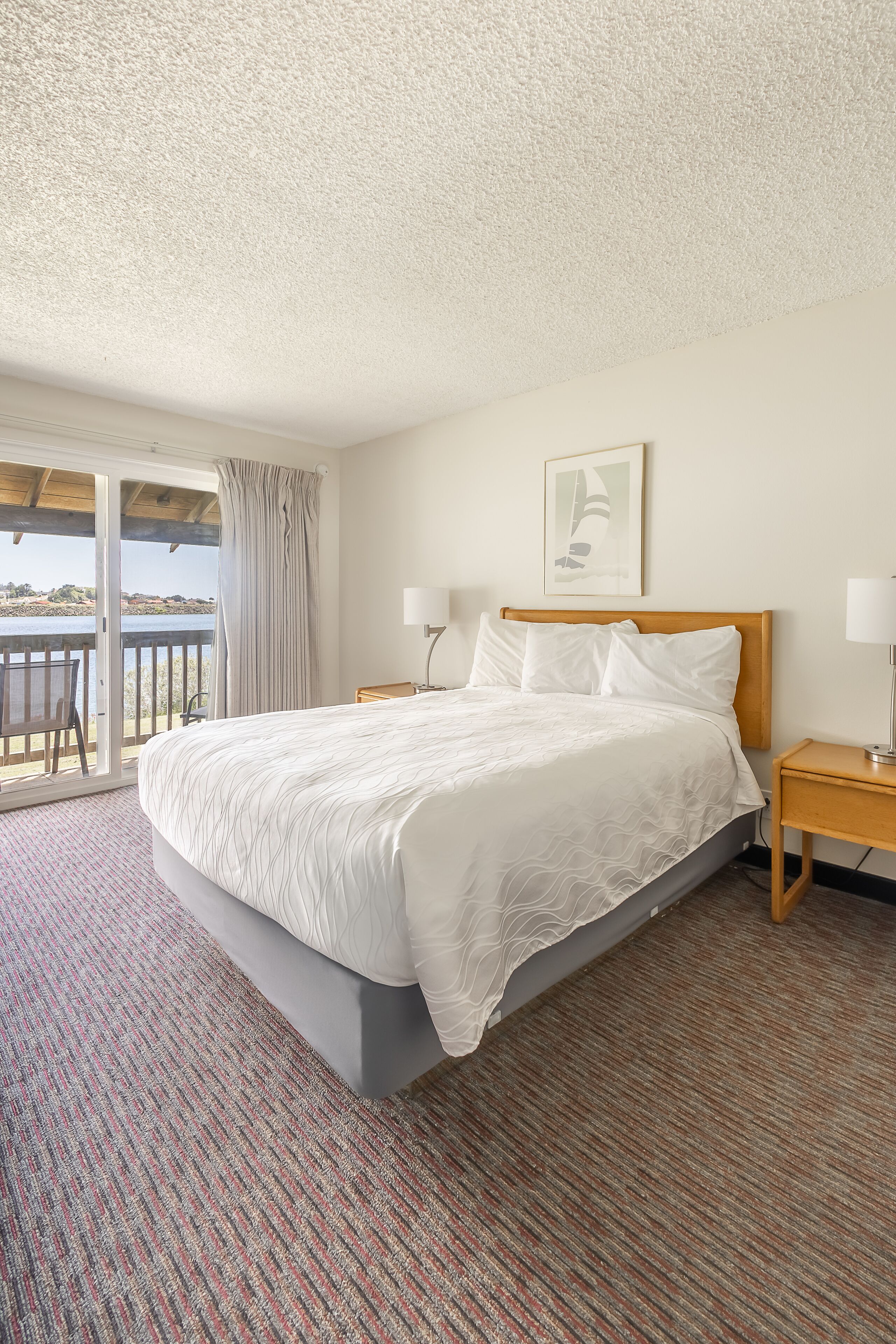 A hotel room with a large bed, a wooden headboard, a nightstand with a lamp, and a view of the outdoors through a sliding door.