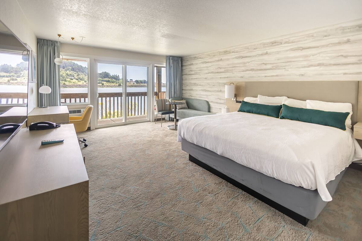 A hotel room with a large bed, a desk with a phone, a chair, and a view of a lake through the balcony doors.