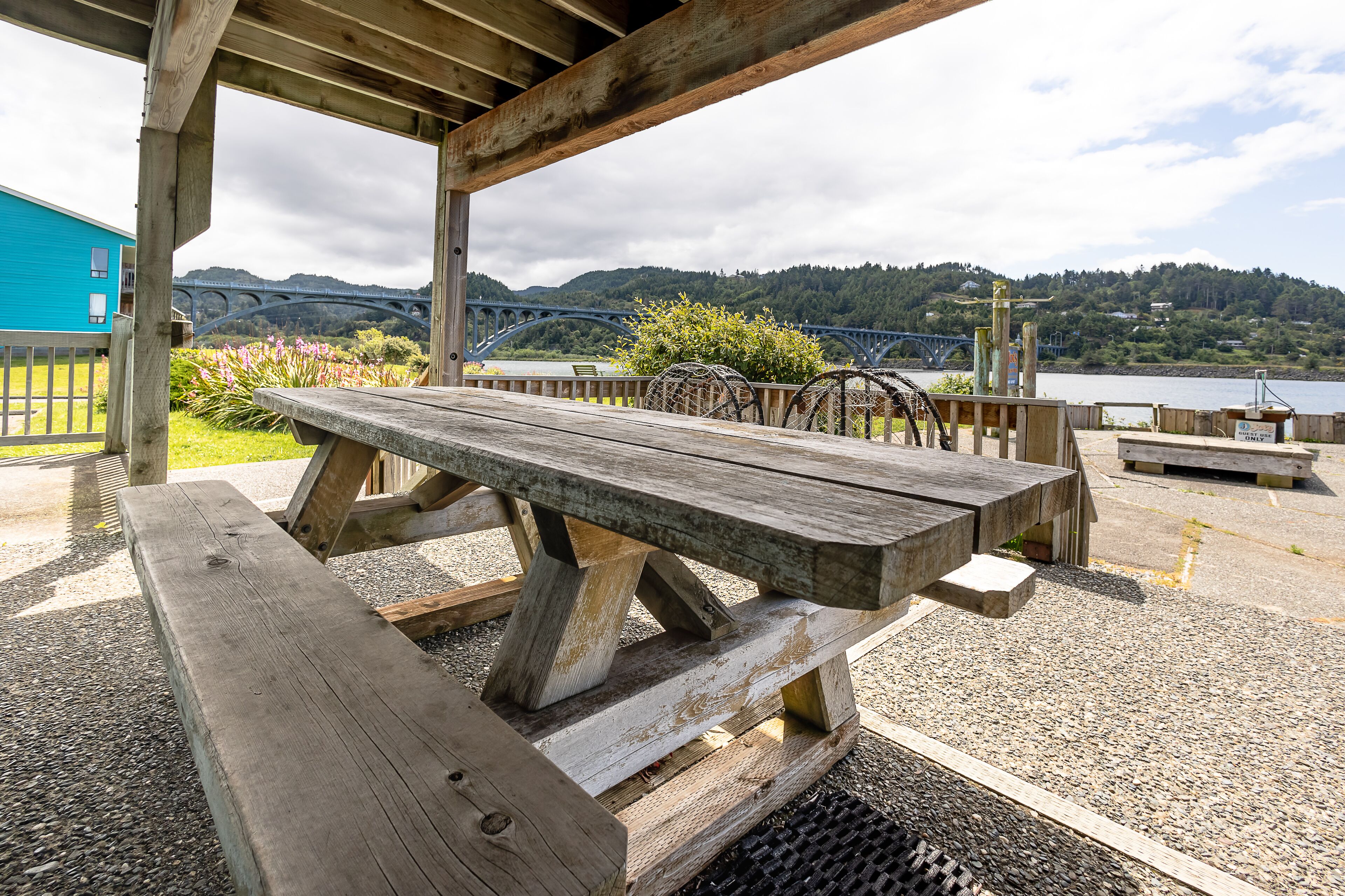Une table de pique-nique en bois, placée sous une pergola en bois, offre une vue sur un pont et des collines en arrière-plan.