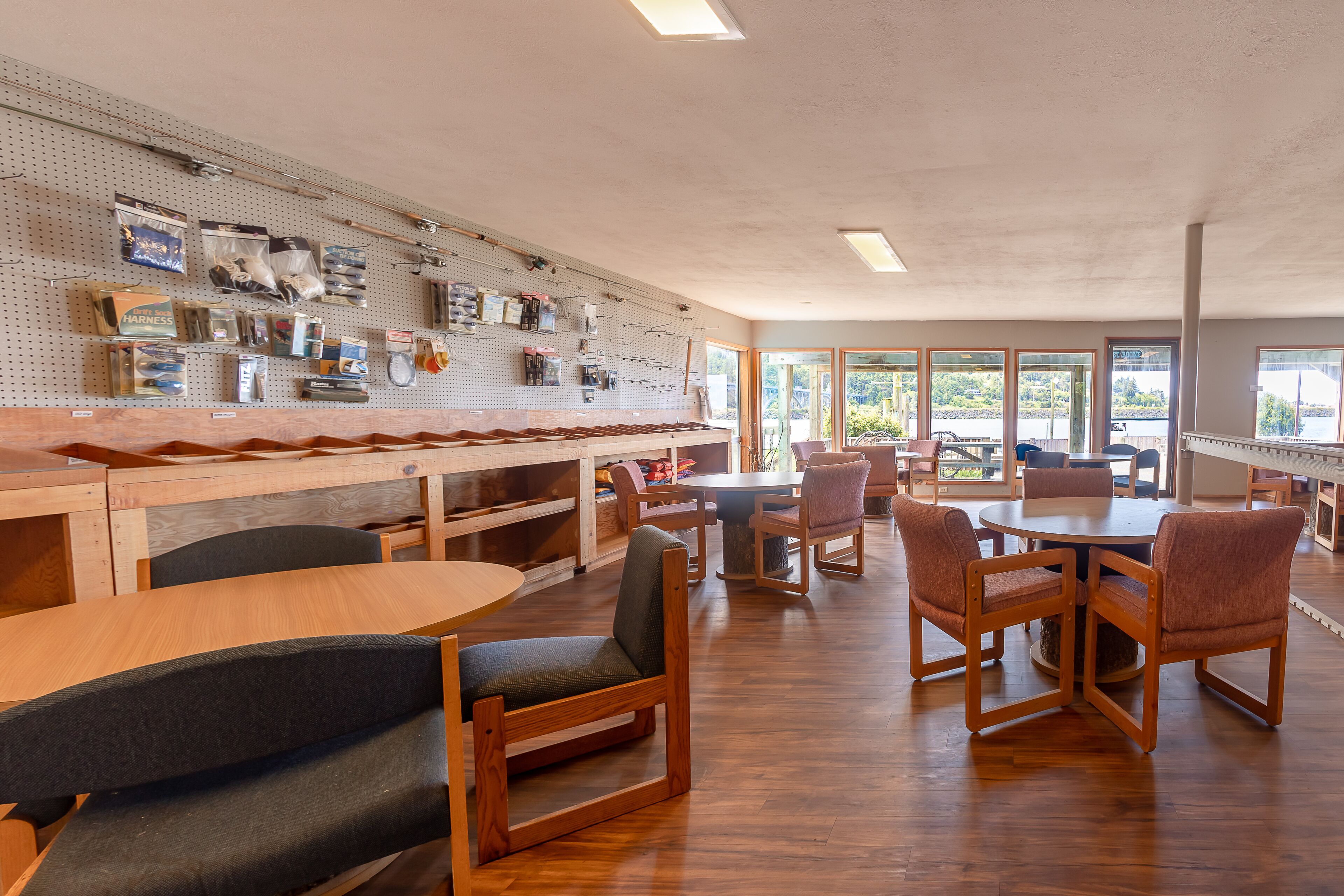 Een eetgedeelte met houten tafels en stoelen, een prikbord met vissersbenodigdheden en grote ramen met uitzicht op de natuur.