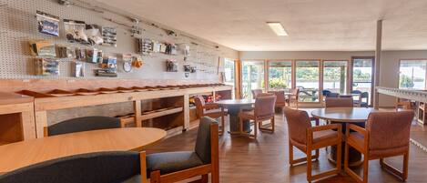 A dining area with wooden tables and chairs, a pegboard wall with fishing equipment, and large windows offering views of the outdoors.