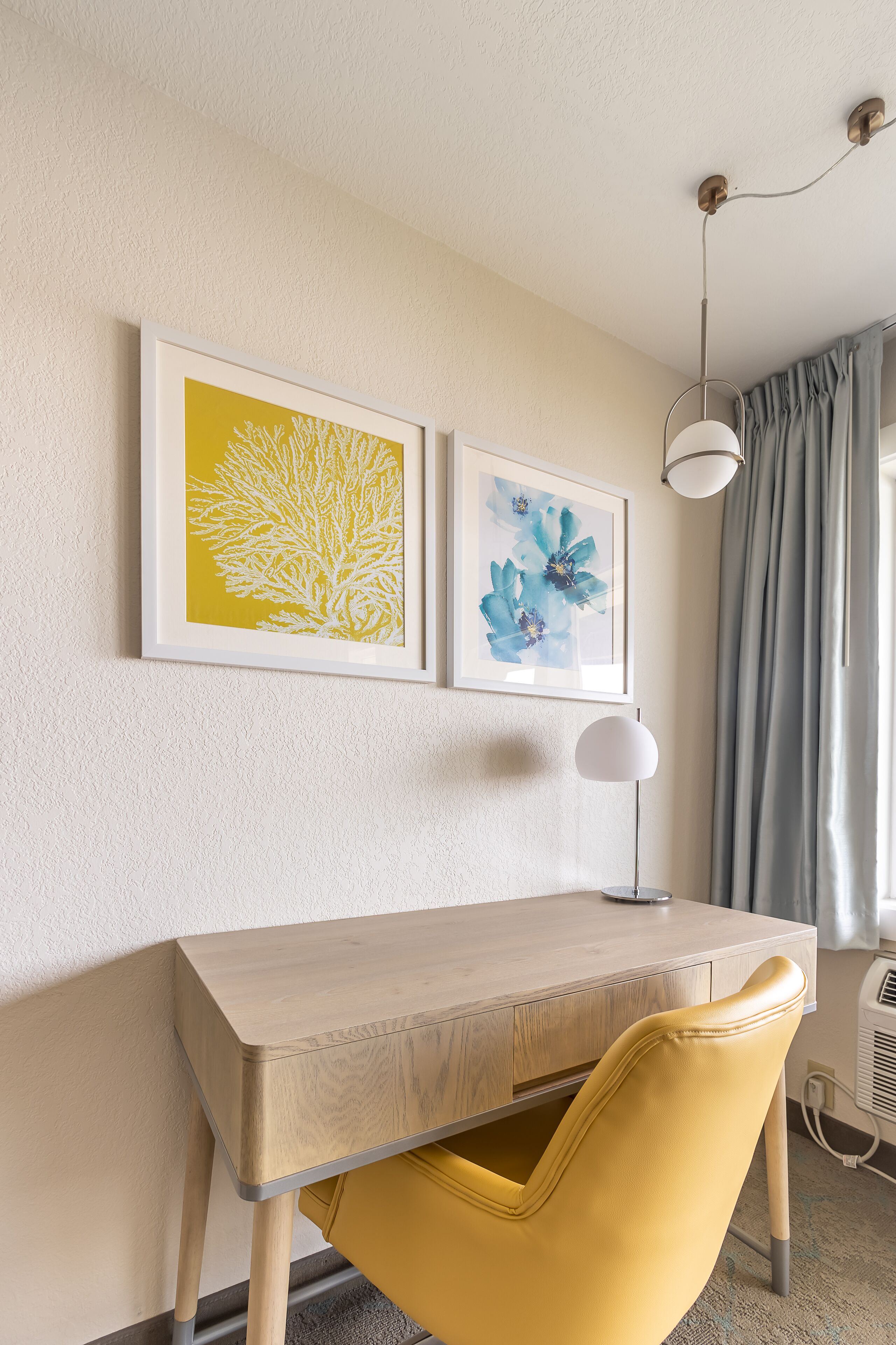 Una habitación con un escritorio de madera, una silla amarilla, dos cuadros enmarcados, una lámpara de pie blanca y una ventana con cortinas.